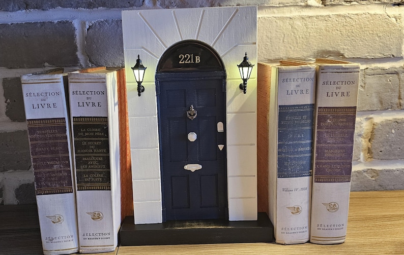 Puede incluir: Una r&eacute;plica en miniatura de una casa adosada de Londres con una puerta azul y el n&uacute;mero 221B sobre la puerta. La casa adosada est&aacute; flanqueada por dos sujetalibros con el texto "Selection du Livre" en los lomos.