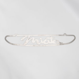 Może przedstawiać: Srebrny metalowy wycięty napis "Miata" z dwoma otworami na górze do zawieszenia.