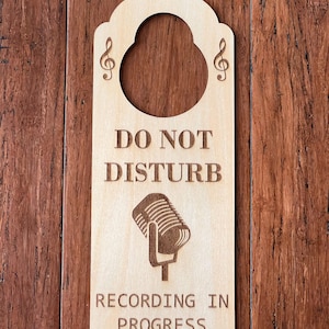 May include: Wooden door hanger with a decorative top and a large circular cutout. The text "DO NOT DISTURB" and "RECORDING IN PROGRESS" are engraved, along with a microphone graphic. Treble clefs are on each side of the cutout.