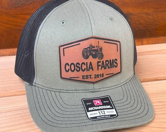 May include: A green and black trucker hat with a brown leather patch that says "Coscia Farms Est. 2016" and a small tractor image. The hat is adjustable and has a Richardson logo.
