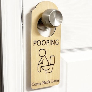 May include: A wooden door hanger with the word "POOPING" above a stick figure on a toilet. The text "Come Back Later" is below the illustration. The hanger is attached to a silver door handle on a white door.