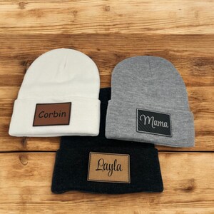 May include: Three knit beanies with personalized leather patches. The beanies are white, gray, and black. The patches have the names "Corbin", "Mama", and "Layla" written in cursive.