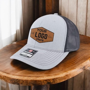 May include: A gray and black trucker hat with a brown leather patch that says "YOUR LOGO Here" and "LK Co."