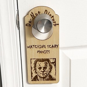 May include: Wooden door hanger with the text "Do Not Disturb" and "Watching Scary Movies". The hanger features a black and white illustration of a scary movie character.