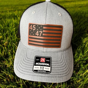 May include: A gray and black trucker hat with a brown leather patch featuring a red, white, and blue American flag with the numbers "45" and "47" on it. The hat has a black mesh back and a label that reads "Richardson. Original 112 Trucker Adjustable."