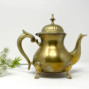 Puede incluir: Una tetera de latón vintage con cuerpo redondeado, asa curva y boquilla. La tetera tiene una tapa con un adorno decorativo y descansa sobre tres pequeños pies. El latón tiene una superficie pulida y reflectante. Una pieza clásica.
