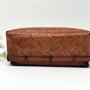 May include: A rectangular, woven basket with a lid. The basket is made of brown bamboo and has a tightly woven, herringbone pattern. The basket is a natural colour and has a simple, elegant design.