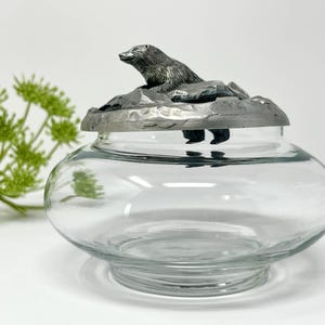 Op de afbeelding: Een heldere glazen pot met een decoratief deksel met een gedetailleerde zilverkleurige beer bovenop een rotsachtig oppervlak. De pot heeft een afgeronde vorm en een brede basis, geschikt voor opslag of weergave.