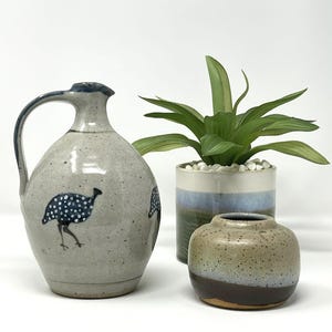 May include: A stoneware jug with a handle, decorated with a blue rim and a hand-painted bird motif with blue spots. A small, round vase and a potted plant with green leaves are also visible.