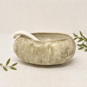 May include: A round, low-profile ceramic bowl with a speckled, off-white and beige finish. The exterior surface features subtle, vertical indentations along the sides. A matching, curved ceramic spoon rests inside the bowl, extending over the rim.