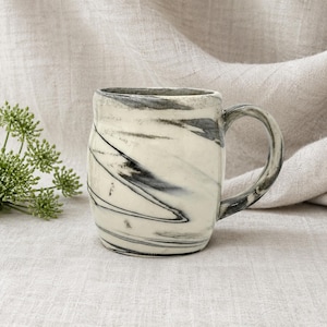 Può includere: Una tazza in ceramica con base bianca e un motivo a spirale nero e grigio. La tazza ha un corpo arrotondato, un manico robusto e una superficie leggermente strutturata. Adatta per bevande calde o fredde.