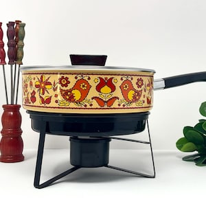 Könnte beinhalten: Vintage Fondue-Topf mit schwarzem Sockel und Griff. Der Topf hat eine cremefarbene Außenseite mit einem Blumen- und Vogeldesign in Rot, Orange und Gelb. Inklusive Fonduegabeln und einem Holzhalter.