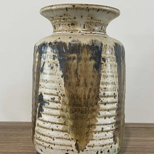 Puede incluir: Un jarrón de cerámica con un cuerpo cilíndrico ancho y un borde acampanado. El jarrón presenta una superficie texturizada con una base color crema y un esmalte goteado marrón y negro. El diseño incluye crestas horizontales y vetas verticales.