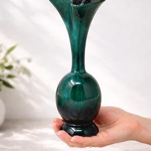 Könnte beinhalten: Eine glänzende, smaragdgrüne Keramikvase mit einem ausgestellten, gerüschten Oberteil. Die Vase hat einen bauchigen Sockel und einen schlanken Hals, der etwa 20 cm hoch ist. Die dunkelgrüne Farbe verleiht ihr eine Vintage-Ästhetik.