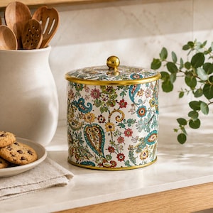 May include: A cylindrical metal canister featuring a white background with a colourful floral and paisley pattern in shades of teal, red, yellow, and green. The canister has a gold-toned rim and a matching gold-toned spherical knob on the lid.
