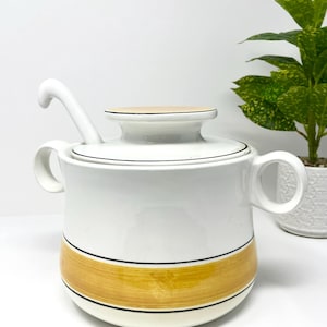 May include: White ceramic soup tureen with a lid and two handles. The tureen has a gold band with black trim and a matching ladle. A small plant is visible in the background.
