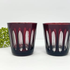 Puede incluir: Dos vasos a juego hechos de vidrio rojo intenso. Cada vaso presenta una serie de cortes verticales ovalados y transparentes en el exterior, creando un patrón repetitivo que revela el vidrio transparente bajo la superficie de color rojo oscuro.