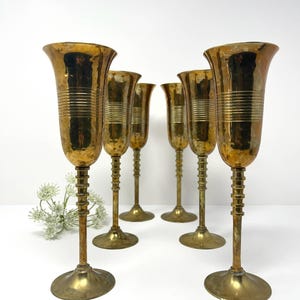 Puede incluir: Juego de seis copas de champán vintage doradas. Las copas tienen un aspecto texturizado y martillado. Cada copa presenta una copa acampanada, un tallo acanalado y una base redonda. Las copas están dispuestas sobre una superficie blanca.