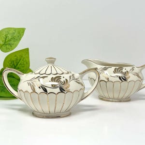 May include: A cream-coloured ceramic sugar bowl with a lid and a matching creamer pitcher. Both pieces feature a fluted base design and are decorated with metallic gold floral patterns and gold trim along the handles and rims.