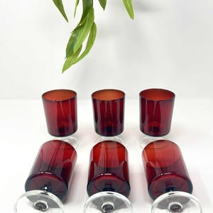 Könnte beinhalten: Sechs rote Weingläser mit klaren Stielen und Böden sind auf einer weißen Oberfläche angeordnet. Drei Gläser stehen aufrecht, und drei liegen auf der Seite. Die Gläser haben eine tiefrote Farbe.