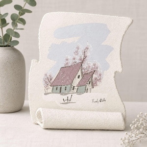 Peut inclure: Une plaque en pierre texturée de couleur blanc cassé, de forme irrégulière, présentant une illustration peinte d'une maison au toit violet et aux murs verts, sur fond de ciel bleu clair et d'arbres roses. La signature est visible en bas à droite.