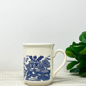 May include: Cream-colored ceramic mug with a blue Willow pattern design. The design features a pagoda, trees, and a bridge. The mug has a curved handle and a slightly flared rim. The mug is sitting on a textured surface.