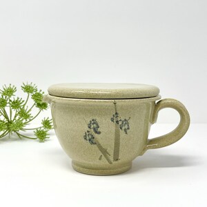 May include: A light beige ceramic mug with a matching lid and a curved handle. The mug features a speckled texture and a hand-painted blue floral design on the front. The piece has a wide, rounded body and a small spout on the left side.