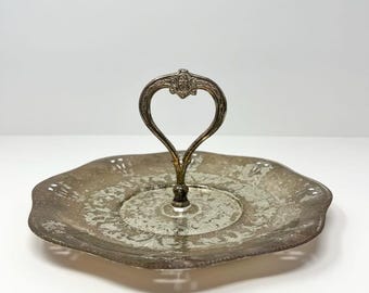 Puede incluir: Una bandeja de servir plateada vintage con un asa en forma de corazón. La bandeja tiene un borde festoneado y un diseño ornamentado. El asa está unida al centro de la bandeja.