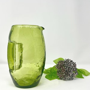 May include: A green glass jug with a textured, crackled surface. The jug has a handle and a spout. A decorative floral arrangement with green leaves and small, dark berries is next to the jug.