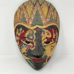 Könnte beinhalten: Eine handgeschnitzte Holzmaske mit komplexen Batik-Mustern in Rot, Gelb, Grün und Schwarz. Die Maske zeigt mandelförmige Augen, eine spitze Nase und rot bemalte Lippen, wobei der obere Bereich eine texturierte, geometrische Gestaltung aufweist.