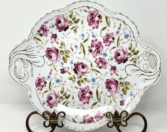 May include: A white porcelain serving tray with a floral pattern of pink roses, green leaves, and blue accents. The tray has a scalloped edge with gold trim and handles. It is displayed on a decorative metal stand.