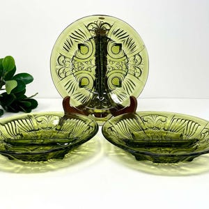 May include: Three olive green glass dishes featuring a pressed pattern of radiating lines and central floral motifs. One dish stands upright on a wooden stand, while two matching shallow dishes with side handles are positioned in the foreground.