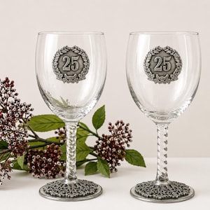 May include: Two clear wine glasses with decorative silver bases and stems. Each glass features a silver emblem with the number 25 inside a laurel wreath. The glasses are displayed with greenery in the background.
