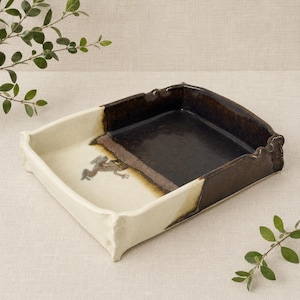 Peut inclure: Un plateau rectangulaire en céramique avec un design bicolore. Un côté est blanc cassé crème avec une petite illustration détaillée d'une grenouille, tandis que l'autre côté est brun foncé profond. Les bords du plateau ont une texture ondulée.