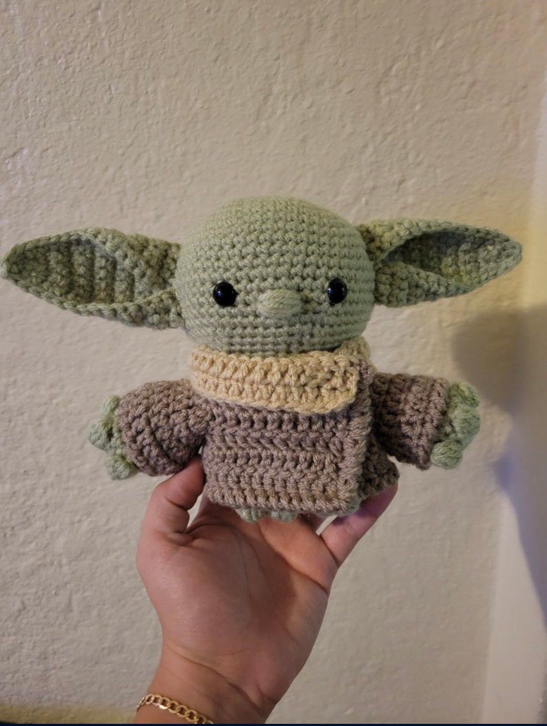 Puede incluir: Un juguete de peluche verde y marr&oacute;n tejido a crochet con orejas grandes y una bufanda. El juguete es un personaje de la franquicia Star Wars.