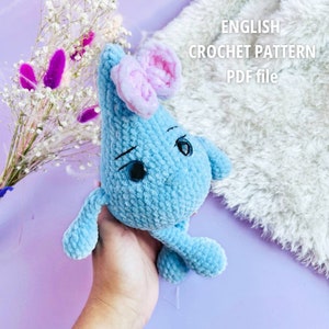Può includere: Un personaggio a forma di goccia di pioggia in crochet blu con orecchie rosa e occhi neri. Il personaggio è tenuto da una mano. Il testo "ENGLISH CROCHET PATTERN PDF file" è visibile sullo sfondo.