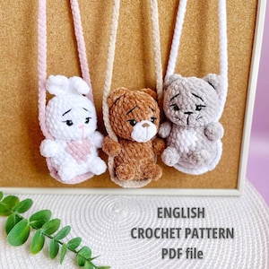 May include: Three handmade crochet plushies: a white bunny with a pink heart, a brown bear, and a gray cat. Each plushie has a strap and is hanging. The text "ENGLISH CROCHET PATTERN PDF file" is at the bottom.