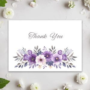 May include: A white card with the words "Thank You" in elegant script. The card features a watercolor floral design with purple, lavender, and white flowers, and gray-green leaves. The card is surrounded by white flowers and green leaves.