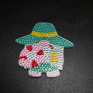 May include: A turquoise, pink, and yellow house-shaped ornament with a hat, made of rhinestones.