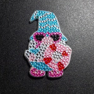 以下が含まれることがあります： 青い帽子とサングラスをかけた、青、ピンク、白のノーム。ノームはラインストーンでできており、装飾品です。