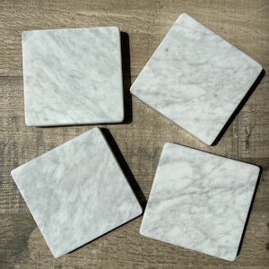 Può includere: Quattro sottobicchieri in marmo bianco con un motivo di venature grigie. I sottobicchieri sono di forma quadrata e hanno una superficie liscia e lucida.