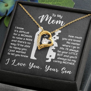 May include: A gold necklace with a heart-shaped pendant featuring a clear crystal. The necklace is in a black box with a message that reads "To My Mom I know it's difficult for a WOMAN to raise a MAN and there's no way I'll be able to repay you. I just wanted to let you know how much you are loved. No matter where I go in life. I'll always be your LITTLE BOY and you'll always be my caring MOTHER. I Love You, Your Son"