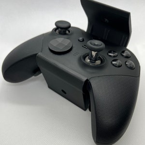 Könnte beinhalten: Ein schwarzer Xbox-Controller mit einer strukturierten Oberfläche und einem schwarzen Telefonständer, der oben angebracht ist. Der Ständer ist geöffnet und der Controller ist zur Kamera gerichtet.