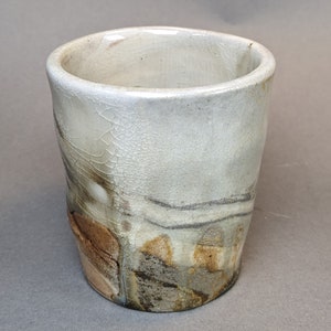 May include: A ceramic cup with a cylindrical shape and a wide opening. The exterior features a crackled white glaze finish that transitions into earthy brown, gray, and charcoal tones near the base, showcasing a textured and uneven surface.