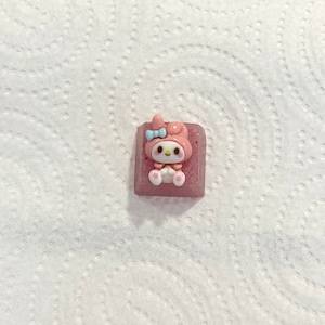 May include: Pink and white keycap with a cartoon character wearing a blue bow. The character has pink ears and a white face with black eyes and a yellow mouth.