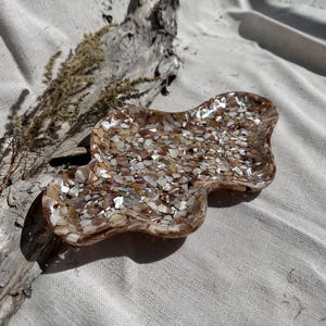 May include: A decorative, free-form dish made of small, iridescent shell pieces. The dish is a mix of beige, brown, and white tones, with a glossy finish. The dish is approximately 6 inches long and 5 inches wide. The dish is on a neutral-colored surface.