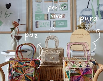 May include: Three handmade woven handbags with handles, each featuring unique colorful thread designs. The bags are displayed on a wicker shelf, with framed art prints in the background. The bags have a rectangular shape.