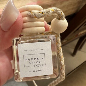 May include: A clear glass bottle with a white label that says "Pumpkin Spice" and a gold and white braided string with a wooden bead. The bottle is 0.33 oz (10 ml).