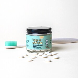 May include: A glass jar with a black lid filled with white toothpaste tablets. The jar has a blue and white label that says "Take us with you" and "Toothpaste Tablets". The label also says "Good for: Festivals, Long Flights, Close Talkers and More". There are also white tablets scattered on a white surface.