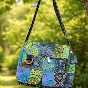 May include: A patchwork messenger bag in shades of blue, green, and purple, with a patterned design. The bag features a flap closure, a zipped pocket, and a wooden D-ring. The strap is black with a patterned design.
